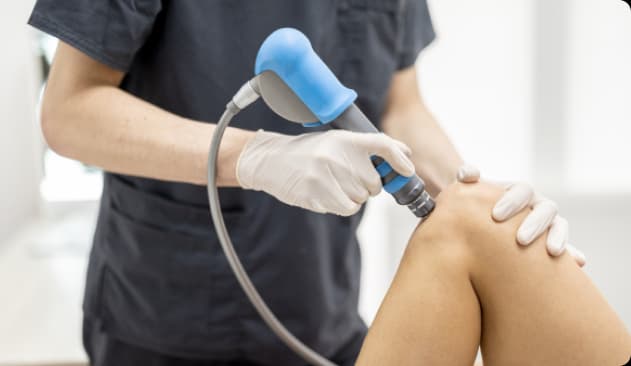 Joelho sendo avaliado a partir da terapia por onda de choque. O aparelho com laterais circulares está sendo segurado por um profissional com luvas. A imagem mostra apenas o joelho, o aparelho e as mãos do especialista.
